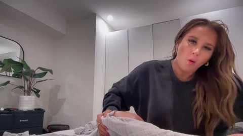 Media: Video of a woman with long brown hair, wearing a black sweater, sitting on a bed, yawning, in a modern, minimalist bedroom with white walls, a potted plant, and a mirror.