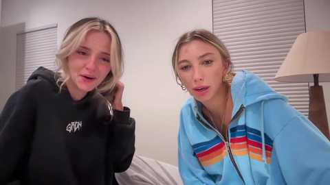 Media: Video of two young women, one blonde in a black hoodie, the other in a colorful striped hoodie, sitting on a bed in a modern, beige room with blinds.