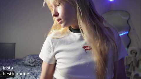 Media: Video of a young blonde girl with long hair, wearing a white t-shirt, standing indoors near a bed with blue sheets and a white lamp in the background.