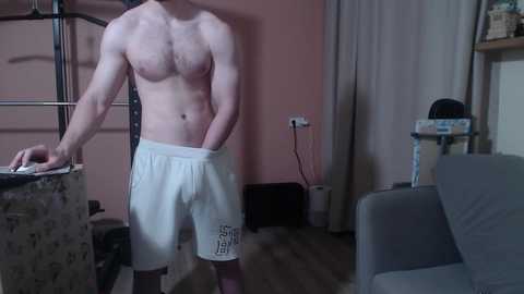 Media: Video of a shirtless, muscular man with pale skin wearing white athletic shorts standing in a cluttered living room with a gray couch, cat tower, and laundry basket.