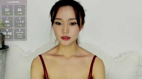 Media: Video of a young East Asian woman with fair skin, dark hair tied back, wearing a red bralette, in a modern, minimalist room with a white couch and a wall chart displaying temperature and humidity levels.