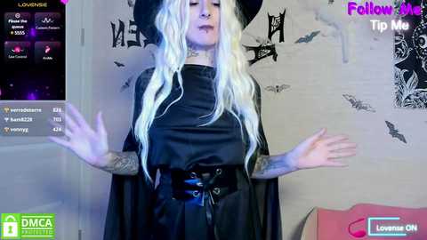 Media: Video of a pale, tattooed woman with long, wavy platinum blonde hair in a black witch costume, with a large hat and wide sleeves. She stands in a room with a Halloween-themed background.