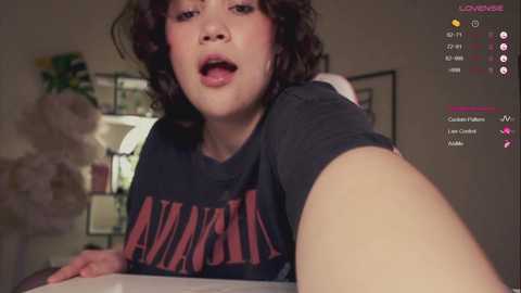 Media: A video of a young person with curly hair, fair skin, and a neutral expression, wearing a black \"Aphasia\" t-shirt, leaning forward. The background shows a blurred indoor scene with a plant and a wall.