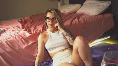 Media: Video of a tattooed, glasses-wearing woman in white crop top and shorts, sitting on a bed with pink and purple bedding, in a cozy bedroom.