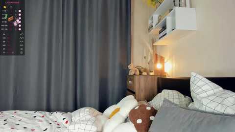 Media: Video of a cozy bedroom with grey curtains, white bed with plaid pillowcases, wooden nightstand holding a lamp and stuffed toys, and a mounted calendar.