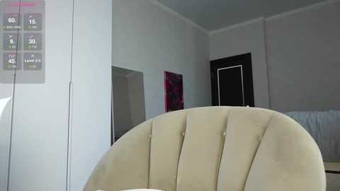 Media: Video of a minimalist room with a beige, tufted armchair, a gray wall, and a closed door with a red patterned wall hanging.