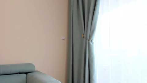 Media: Video of a minimalist room with a light green couch against a pale beige wall, featuring a sheer white curtain tied with a golden tassel, allowing soft natural light to filter through.