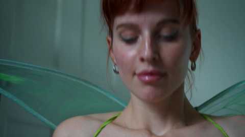 Media: Video of a fair-skinned woman with red hair, wearing a green dress and delicate fairy wings, standing against a muted background.