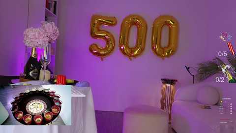 Media: Video of a party room with 500 gold balloons on a white wall, champagne bottles, flowers, and a poker table, purple lighting.