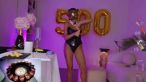 Media: Video of a slender, light-skinned woman in a black bikini, with a gold mask, posing in a purple-lit room with gold balloons spelling \"SOON.\