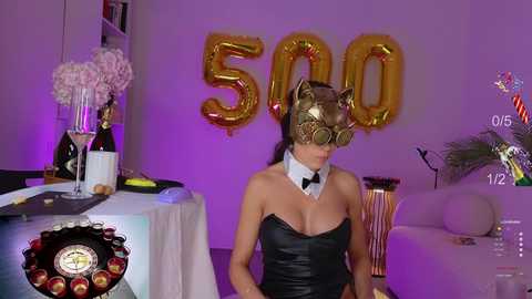 Media: Video of a woman in a black leather strapless dress, gold cat mask, and white bow tie, standing in a dimly lit room with purple lighting, decorated with balloons and champagne bottles, celebrating 500 people.