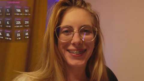 Media: Video of a smiling, fair-skinned woman with long blonde hair, wearing glasses and a black top, standing indoors next to a wooden door with a calendar displaying dates and temperatures.