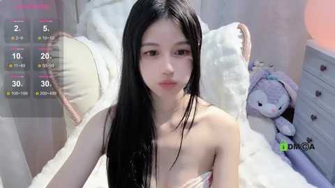 Media: A video of an East Asian woman with long black hair and fair skin, wearing a strapless dress, sitting on a bed in a pastel-themed room.