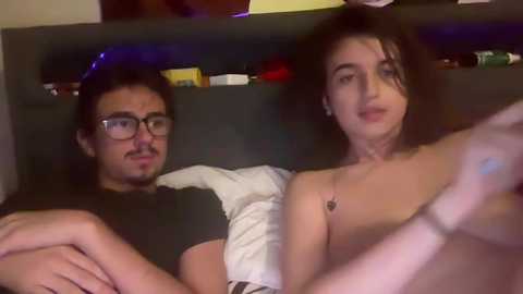 Media: Video of a young, dark-haired woman with medium skin, topless, and a man with glasses and a mustache, both lying on a bed, dimly lit room in the background.