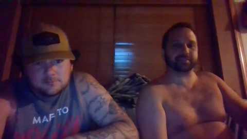 Media: Video of two shirtless men, one with tattoos, wearing a baseball cap, the other with a beard, sitting indoors; wooden wardrobe in background, dim lighting.