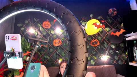 Media: A video of a Halloween-themed room with a large tire mounted on a wall, decorated with jack-o'-lanterns, bats, and a pumpkin. A smartphone with a white case is held against the tire, capturing the scene.