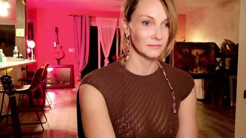 Media: Video of a blonde woman with fair skin and medium build, wearing a brown sequin top, standing in a brightly lit, modern bedroom with pink accents and a vanity table.