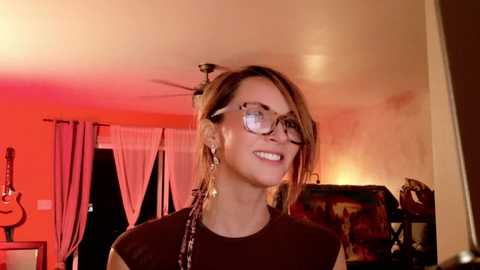 Media: Video of a smiling woman with glasses, wearing a black top, in a warmly lit living room with orange ambient lighting, visible through sheer curtains.