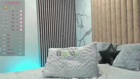 Media: Video of a modern bedroom with a quilted light gray bedspread, black leather pillows, and vertical black-striped wallpaper. A digital calendar display shows upcoming dates and events.