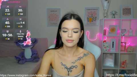 Media: Video of a fair-skinned woman with long black hair, wearing a grey tank top, sitting in a room with neon pink lights, cat statue, and music notes overlay.