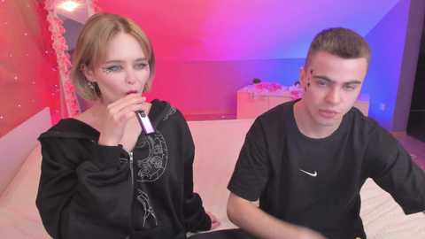 Media: Video of a blonde woman with short hair in a black hoodie and a young man with short hair in a black t-shirt, both holding a purple lipstick. Background is pink and blue with white bedding and a bed.