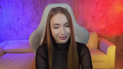 Media: Video of a smiling young woman with long, straight brown hair, wearing a sheer black top, seated on a white gaming chair, with a colorful, abstract background featuring purple, blue, and red hues.