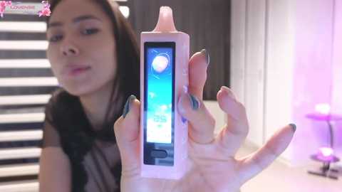 Media: Video of a young Asian woman with long black hair, holding a white rectangular phone with a blue screen, in a modern, well-lit room with vertical blinds and purple lamps.