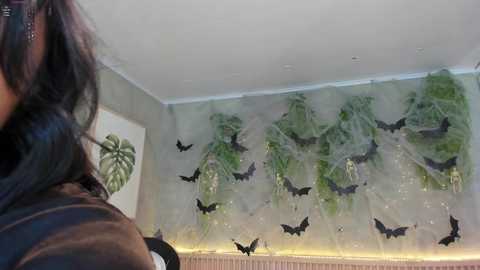 Media: Video of a person's shoulder with dark hair, standing in a room with a mural of bats and greenery on a light-colored wall.