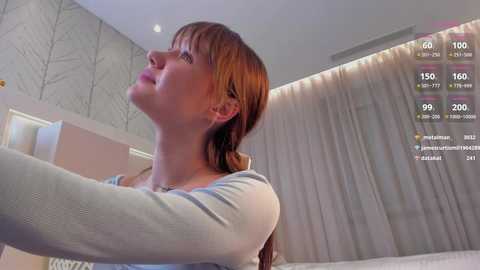 Media: A video of a fair-skinned woman with auburn hair in a light gray top, captured from a smartphone's first-person perspective, standing in a modern bedroom with beige walls and a large window.