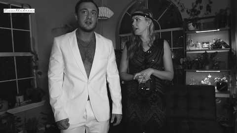 Media: Black-and-white video of a man in a white suit and a woman in a patterned dress, both with serious expressions, standing in a dimly lit living room with shelves filled with decor and a window in the background.