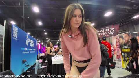 Media: Video of a young woman with long brown hair in a pink hoodie and black shorts, leaning on a table at an indoor event with banners and attendees.