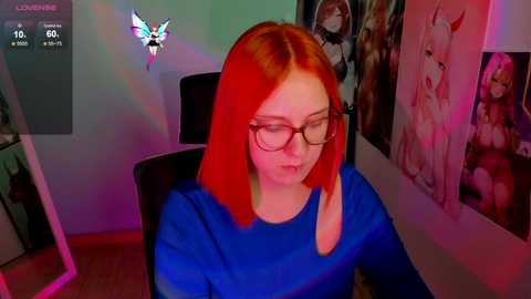 Media: Video of a fair-skinned woman with red hair, wearing glasses, a blue top, and sitting in a dimly lit room with anime posters on the wall.