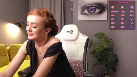 Media: Video of a smiling, fair-skinned woman with red hair, wearing a black off-shoulder dress, sitting on a yellow couch in a modern, gray-toned room with a large white gaming chair, a digital monitor, and a potted plant.