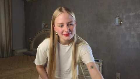 Media: Video of a young blonde woman with straight hair and fair skin, wearing a white t-shirt, sitting in a dimly lit, gray-toned room with a wooden floor and a decorative fireplace in the background.