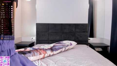 Media: A video of a modern, minimalist bedroom with a large bed featuring a black tufted headboard, white and purple striped bedspread, and a pillow with a floral pattern. The walls are light gray, and there are two black bedside tables.