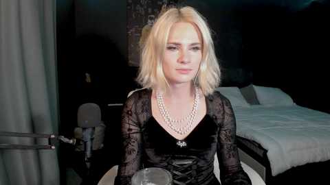 Media: A video of a blonde woman with shoulder-length hair, wearing a black velvet dress with lace sleeves and a silver necklace, sitting at a round table in a dimly lit bedroom with a white bed and dark walls.