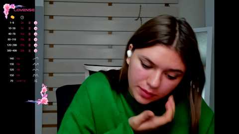 Media: Video of a young woman with fair skin and straight brown hair, wearing a green sweater, talking on a white earpiece. Background shows a white wall with horizontal blinds.