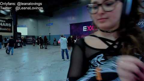 Media: Video of a dimly lit convention hall with a young woman in glasses, a black choker, and a black mesh top, looking at a screen. Background includes attendees and signs.