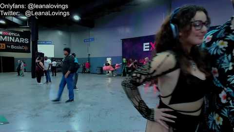 Media: Video of a large, dimly-lit convention center with a tall, slender woman in black lingerie and floral jacket, standing beside a man in a hoodie. Background includes people in casual clothes and a sign reading \"BETA.\