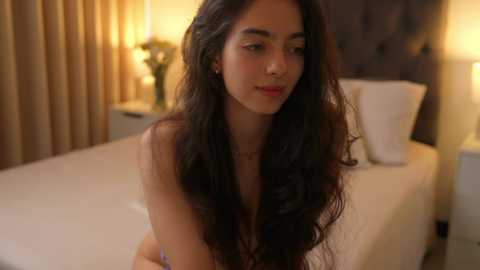 Media: A video of a young woman with long, wavy dark hair, light skin, and a slender physique, sitting nude on a bed with beige sheets in a softly lit room.