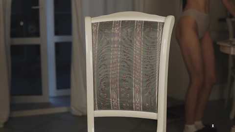 Media: Video of a woman in light grey underwear standing in a dimly lit room, wearing a white headband, with a patterned backrest chair in the foreground.