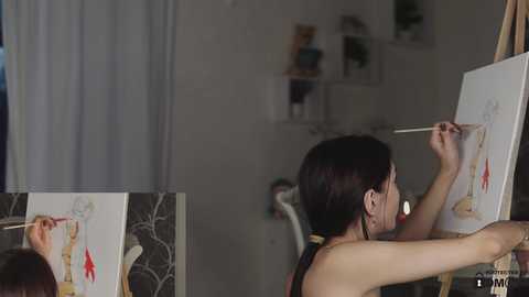 Media: Video of an artist with short dark hair, painting in a studio. She's focused on an anatomical drawing of a human body on a white canvas. The background features a white wall with shelves holding books and plants.