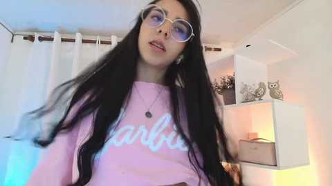 Media: Video of a young woman with long black hair, wearing large round glasses and a pink \"Lamb\" sweatshirt, standing in a modern, white-themed room with shelves, owls, and a lit lamp.