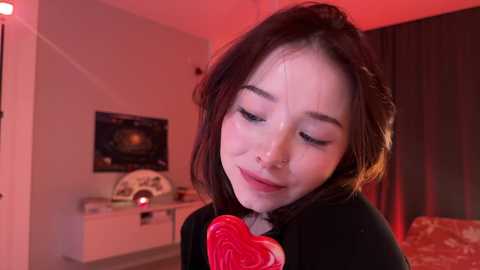 Media: Video of a young Asian woman with fair skin, wearing a black dress with a red rose, smiling, with semen on her face, in a dimly lit, red-lit room with a white dresser and wall art.