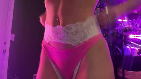 Media: Video of a woman in a low-cut, white lace bralette and pink thong, emphasizing her toned midriff and hips. Background features a dimly lit room with purple lighting and a mix of furniture and objects.