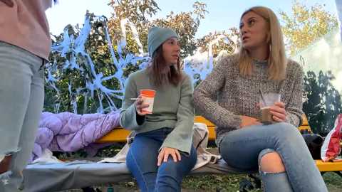 Media: Video of two young women, one in a gray sweater and beanie, the other in a knit sweater, seated on a yellow bench. They hold cups, surrounded by a colorful, festive scene with trees and white decorations.