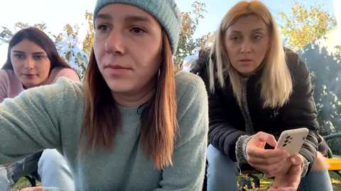 Media: Video of three young women, one in a blue beanie, two with long hair, outdoors in a park, all looking serious.