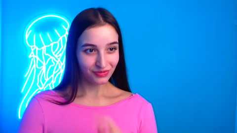 Media: Video of a young woman with long, straight brown hair, fair skin, and a light pink top, standing against a bright blue backdrop with a glowing neon jellyfish outline.