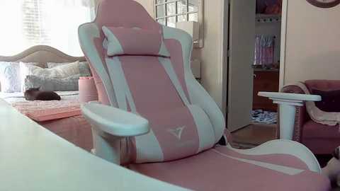 Media: Video of a pink gaming chair with white accents, situated in a cozy, cluttered bedroom with a bed, a cat, and a white cat tree in the background.