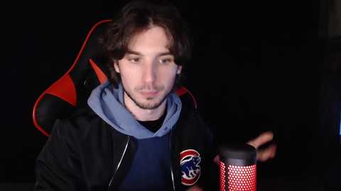 Media: Video of a young man with curly dark hair, light skin, wearing a blue hoodie, red and black gaming chair, holding a black and red headset microphone.